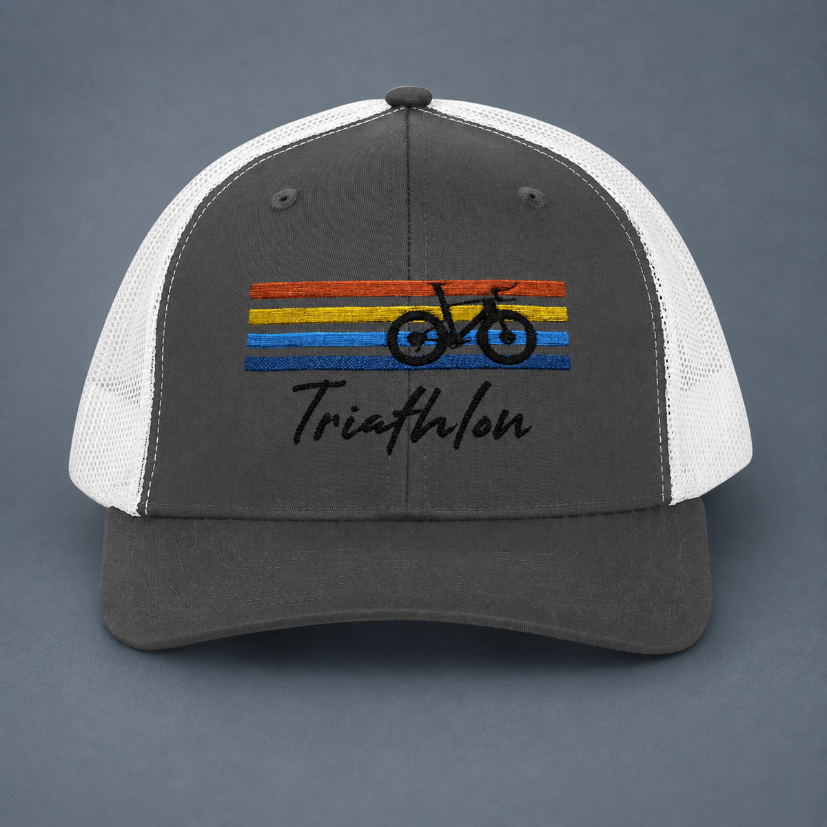 A dark gray and white trucker-style cap with a black embroidered bicycle silhouette over horizontal orange, yellow, and blue stripes on the front panel, plus the word 'Triathlon' in black script below. The cap is photographed against a smooth dark gray background.