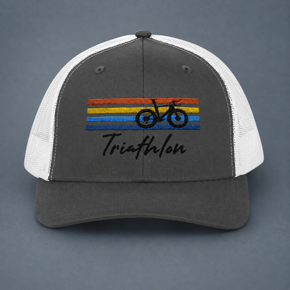 A dark gray and white trucker-style cap with a black embroidered bicycle silhouette over horizontal orange, yellow, and blue stripes on the front panel, plus the word 'Triathlon' in black script below. The cap is photographed against a smooth dark gray background.