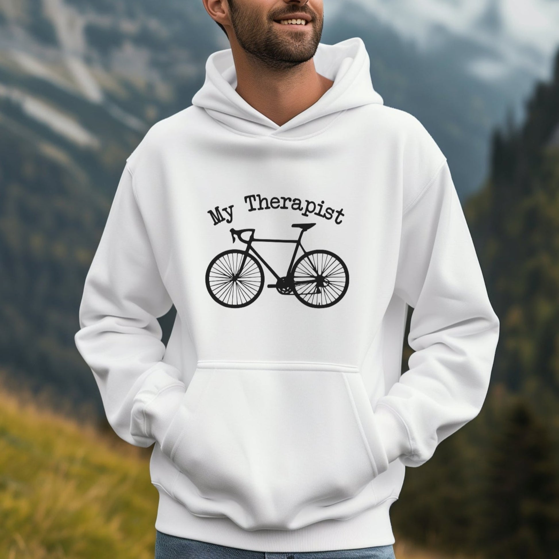 Man wearing a white hoodie with a bicycle graphic and 'My Therapist' text, standing in a mountainous landscape.