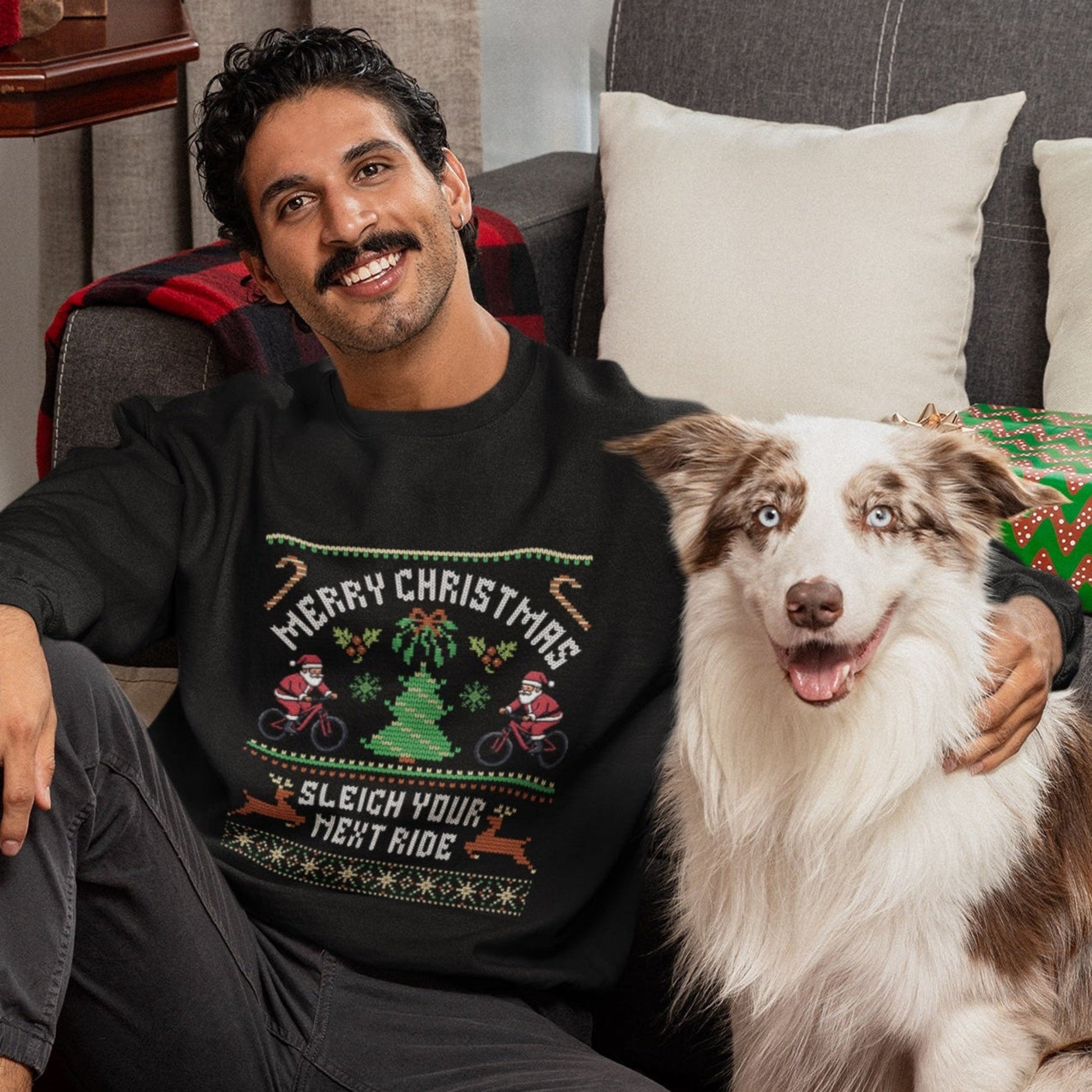 Man wearing a Christmas-themed sweater sitting with a dog in a festive living room.